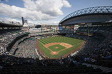 A panoramic view of a baseball stadium with a large crowd and open roof, looking over the diamond from above home plate.