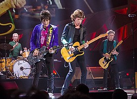 Rolling Stones předvádění na jevišti v Milwaukee ve Wisconsinu. Zleva: Charlie Watts na hnědé bicí soupravě, Ronnie Wood v purpurové bundě s černými džíny hrající na stříbrnou kytaru, Mick Jagger v černé košili a kalhotách na oranžově / žluté kytaru, Keith Richards se zelenou vestou a černým oblečením oranžová / žlutá kytara (podobná Jaggerově)