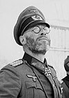 A black-and-white photograph of a man wearing a military uniform e a neck order in shape of an Iron Cross. He is wearing glasses e is looking up into the sky.