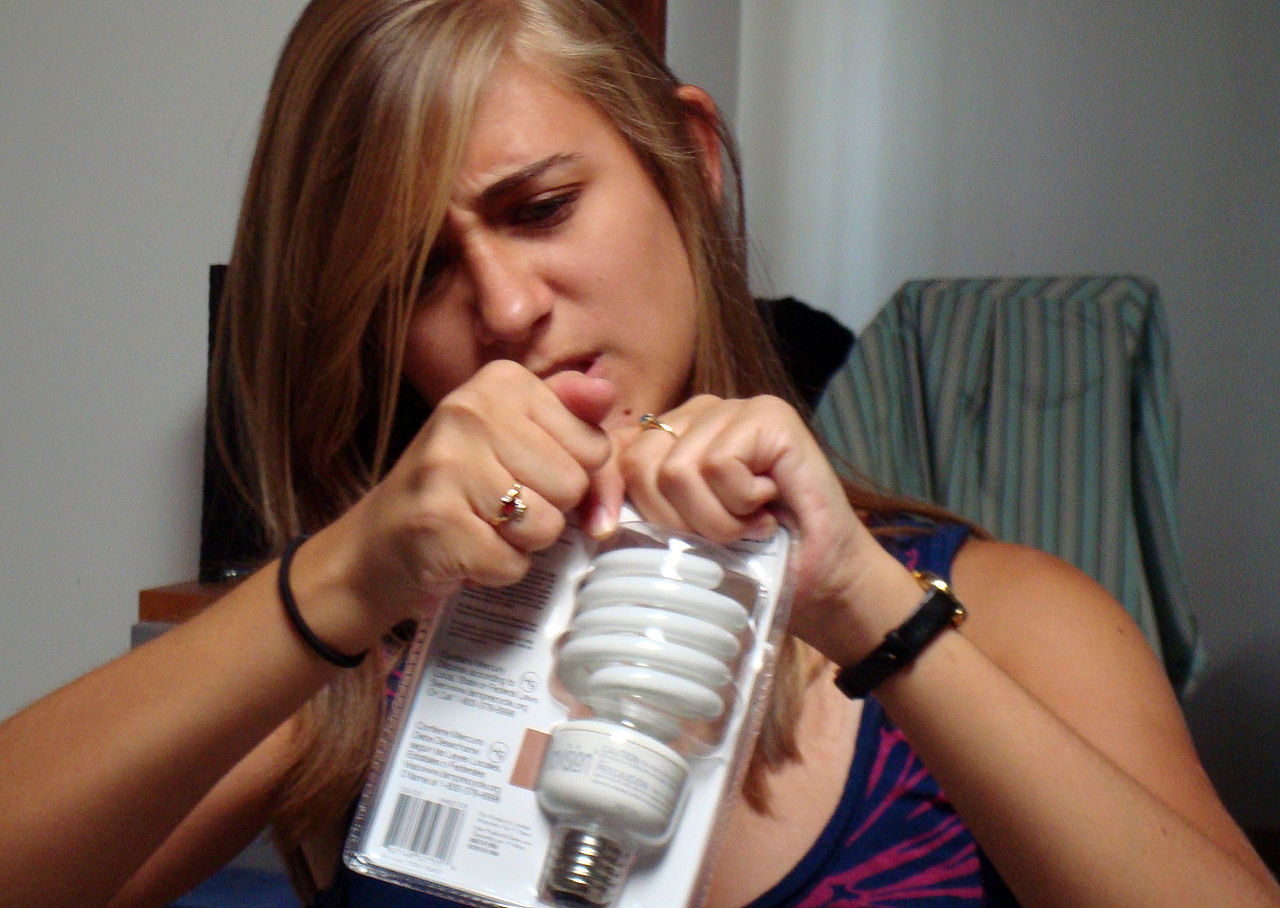 A girl is attempting to open a plastic package containing a light bulb.