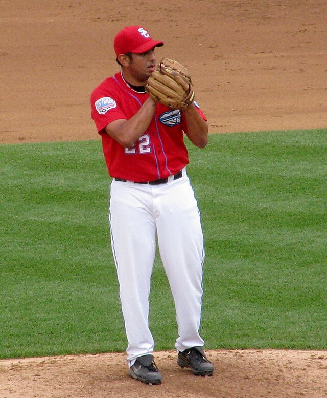 A man wearing a red baseball jersey and cap and white baseball pants standing on a grass mound