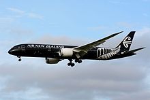 A mid-size jet airliner in flight. The plane livery is all-black and features a New Zealand silver fern mark.