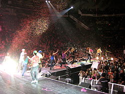 Группа танцоров в ярких одеждах спускается по подиуму под падающим сверху конфетти. В самом конце подиума, рядом с толпой, стоит брюнетка-подросток в розовом наряде.
