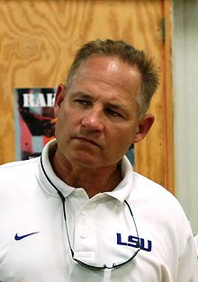 A man wearing a white collared shirt with sunglasses around his neck. His head is cocked to his left looking to his right.