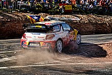 Citroën DS3 WRC de Sébastien Loeb, livrée rouge, bleue et blanche, en sortie d'épingle sur une route goudronnée ; spectateurs en arrière-plan.