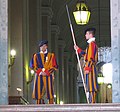 La porte de Bronze, entrée principale du palais apostolique du Vatican, gardée en permanence par deux hallebardiers de la Garde suisse pontificale. A l'intérieur, à gauche, le sergent se tient derrière un bureau. L'Escalier royal est visible au fond, à l'arrière-plan.