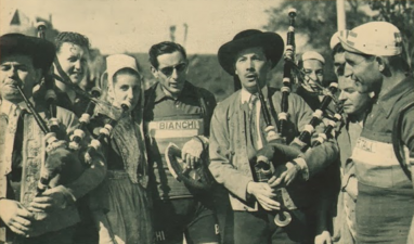 Photo en noir et blanc montrant deux hommes jouant d'un instrument de musique, à leurs côtés des coureurs cyclistes et une femme portant une coiffe blanche