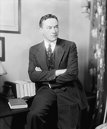 Липман, облечен в костюм, облегнат на бюро със скръстени ръце