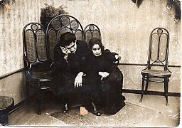 Black and white photo showing a man and a woman, dressed in black, crying, sitting on chairs.