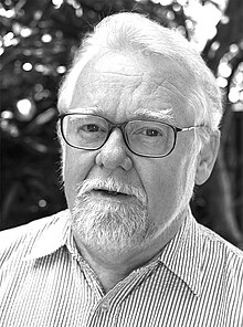 Head and shoulders monochrome portrait of bearded 58-year-old man in narrow glasses, cheap shirt, tieless, with partially opened mouth.