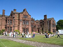 A three-storey house with bays containing mullioned and transomed windows, and multiple gables. On the lawned area in front of the house are many people presumably enjoying an open day.