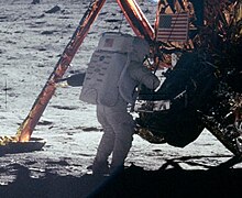 Зернистое изображение сзади человека в белом скафандре и рюкзаке, стоящего перед Лунным модулем на поверхности Луны. Видна посадочная нога и флаг США на спуске.
