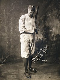 A black-and-white photo of a man posting in front of the camera. He is wearing his uniform, holding a baseball bat. He is posing for the camera but looking away slightly from it. An autograph of the subject is also seen in the middle of the picture.
