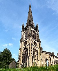 An elaborately decorated tower in light stone with dark stone dressings, windows of varying shapes and sizes, pinnacles, and a spire