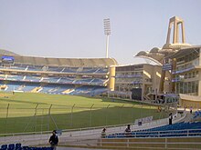Back view of the entrance and a stand of a cricket stadium. Some people can be seen near the seats.