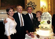 Two men in suits are flanked by two women in formal dresses, standing beside a large birthday cake with lit candles and flowers. The cake is decorated with the text "Happy 90th Birthday President Ford".