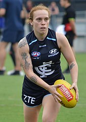 A female athlete in a navy blue sleeveless guernsey running with a football