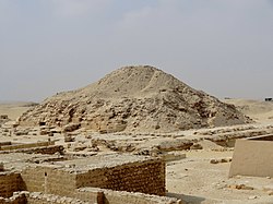 A photograph of the ruined pyramid of Unas. Parts of the base of the pyramid remain generally intact, with visible polished stone blocks, while the head of the pyramid resembles a large mound of sand. The ruins of the mortuary temple can be seen in the foreground. Segments of the bases of the walls of the mortuary temple and pyramid complex have been retained, though much of the structure is in ruins.