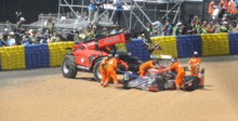 A race car being removed from the circuit after an accident by five people wearing orange for visibility