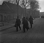 Photo noir et blanc montrant des hommes en uniforme marchant dans une rue. Les deux hommes au premier plan sont désarmés et portent leurs affaires tandis que ceux à l'arrière plan sont armés et les escortent.