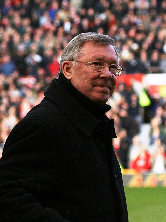A side-on photograph of a man with grey hair wearing glasses and a black overcoat.