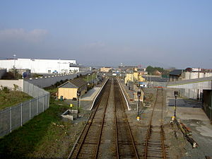 Aerial view of the station taken during the day (with clear blue sky). There are two platforms, with tracks in the centre. On left side, there is the station boundary with a white building on top left corner. The left platform also has a (yellow) shed and a lawn. The right hand side, there is the station office, and parking lot. There is a factory in view. Beyond the platform, there are buildings.