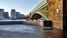 View of an empty paved area that is part of a public riverside footpath, with a railway bridge, next, to the right. Backdrop of the river Thames.