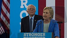 Clinton standing at a podium speaking and looking to her right; Bernie Sanders is standing behind her.