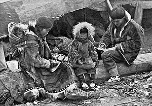 An Inuit family is sitting on a log outside their tent. The parents, wearing warm clothing made of animal skins, are engaged in domestic tasks. Between them sits a toddler, also in skin clothes, staring at the camera. On the mother’s back is a baby in a papoose.