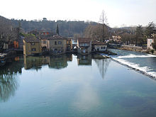 Photo of houses next to a river which flows past in the foreground and right. There is a high bluff on the left side.