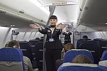 Photo prise à l'intérieur d'une cabine d'un avion. À gauche et à droite de l'image, des sièges accueillent des passagers pour le vol. Au milieu, dans l'allée centrale, deux femmes, dont l'une est à l'avant-plan et l'autre à l'arrière-plan, effectuent des gestes afin d'expliquer les consignes de sécurité.