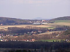 Zdjęcie przedstawia górski krajobraz sfotografowany ze stoku. Kadr wypełniają niskie góry o łagodnych zboczach pokryte lasami, łąkami i polami. W dolinach między pagórkami znajdują się zabudowania wsi - niskie domy z dwuspadowymi dachami. Wśród nich wyróżnia się budynek kościoła z dominującą wieżą.