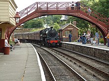 Un train à vapeur rouge et noir arrivant à quai et passant sous une passerelle rouge