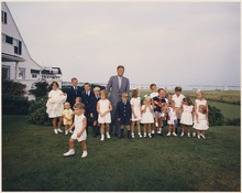 L'ancien président des États-Unis, John F. Kennedy, avec différents enfants de sa famille. Tous se tiennent debout sur la pelouse, derrière laquelle il est possible d'apercevoir une maison blanche ainsi qu'un lac où plusieurs embarcations naviguent.