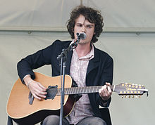 Jon is seated, strumming his acoustic guitar and singing into a microphone