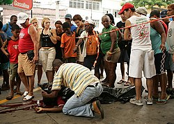 Pai lamentando a morte do filho que está estendido na rua sangrando. Há populares ao redor.