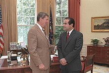 An elderly man in a beige suit is turned profile to the camera and is talking to Scalia, who has his hands folded in front of him as both men stand before an ornate desk.