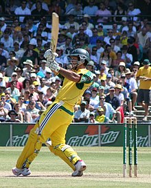 A man in a cricket uniform swinging the bat at a sports ground. A crowd watches in the background.