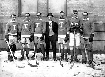 Seven men in row standing in front of brick wall. Six of the seven are wearing hockey sweaters and uniforms