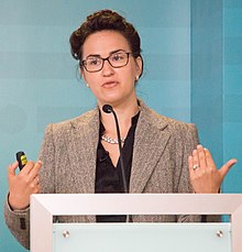 Athena Aktipis wearing a black blouse and a tweed blazer, standing behind a podium, speaking into a microphone, with a clicker in hand