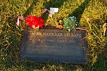 Плоский бронзовый надгробный указатель, окруженный травой, украшенный цветами и маленькими американскими флагами.