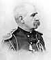 Profile of a balding white man with bushy, drooping mustache wearing an ornate military jacket with shoulder boards, shoulder cords, and a lanyard hanging from the chest.