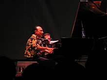 A man in a patterned shirt playing a piano in a dark-lit room. Behind him is a man holding a video camera pointed towards the piano player's hands.