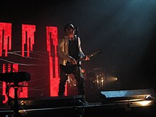 A man playing guitar and singing into a microphone on stage in front of a series of red teeth-like light patterns.