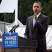 Obama gestures from the podium while campaigning. The front of the podium has a sign that reads "Change We Need" with WWW.BARACKOBAMA.COM below and his campaign logo above.