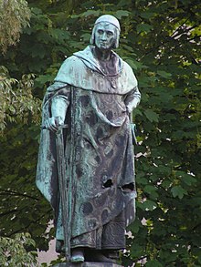 Bronzová socha stojícího může v kněžském rouchu s upřeným pohledem, jež má jednu ruku v bok a v druhé drží meč zabodnutý do podstavce sochy. V pozadí se nachází koruny stromů.