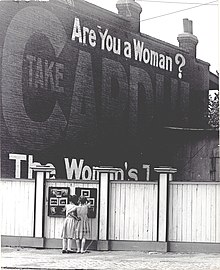 Two girls examining a bulletin board posted on a fence. An advertisement painted above them asks "Are You a Woman?". Two girls examining a bulletin board posted on a fence. An advertisement painted above them asks "Are You a Woman%3F".jpg
