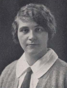 A young white woman with dark hair and eyes, wearing a white shirt with a wide collar and a dark necktie, and a soft cardigan