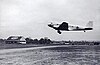 A black and white photograph of a propeller-driven passenger airplane skimming over a highway at a sod airport