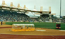 Interior of a sports stadium. There is a running track surrounding a central grassed area. In the distance there are stands full of people